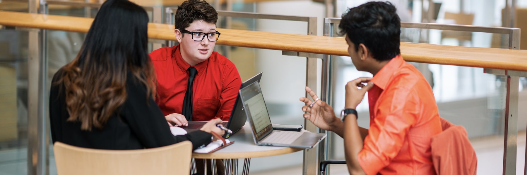 three students sitting around small circular table and laptop one student gesturing with hand