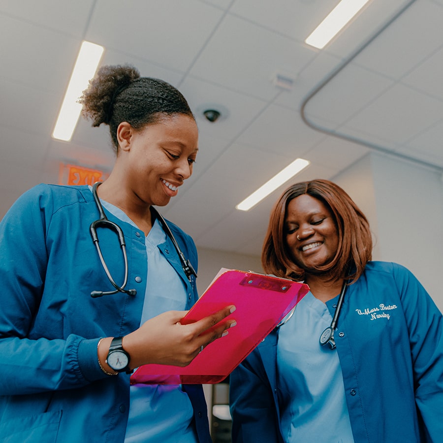 Nursing two students in scrubs hold clipboard in lab.