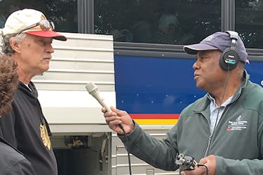 man wearing headphones and carrying recorder conducts interview with man in baseball hat.