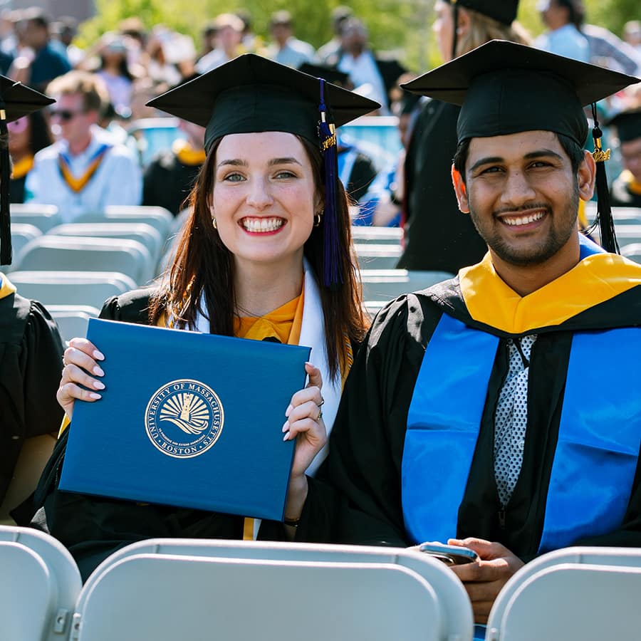 2 sit outside at commencement in caps & gowns, 1 holds up diploma.