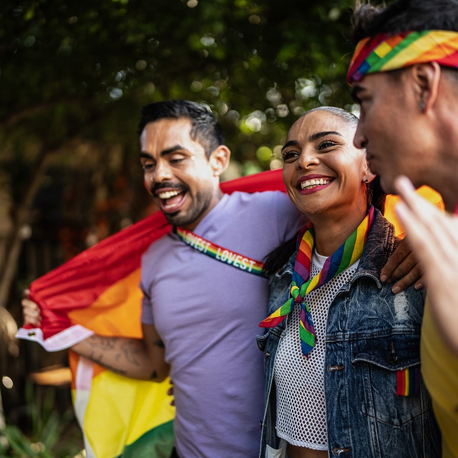 Happy LGBTQIA+ friends talking while walking with a rainbow flag through the city
