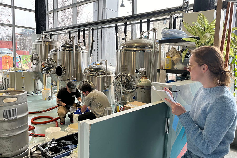 Shelby Ansel, holding a notebook, supervises two men working on brewing equipment in a large, sunlit room. More gleaming vats and tanks line the back wall.